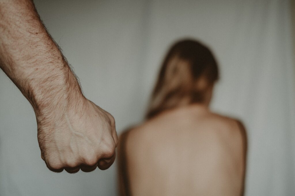 A stock photo implying domestic violence highlighting a foreground with a fist and a blurry woman cowering facing away
